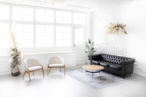 東京都内10施設レンタル撮影スタジオ「GARDEN STUDIO（ガーデンスタジオ）」のSTUDIO TWOBEE（スタジオ トゥービー）の内装写真。大きな窓から光が差し込む真っ白な壁の室内に、黒いレザーのソファー、ファブリックと木材でできたお洒落な椅子が2脚、明るい木製のテーブルが配置されている。落ち着いた雰囲気の観葉植物やスワッグが飾られている